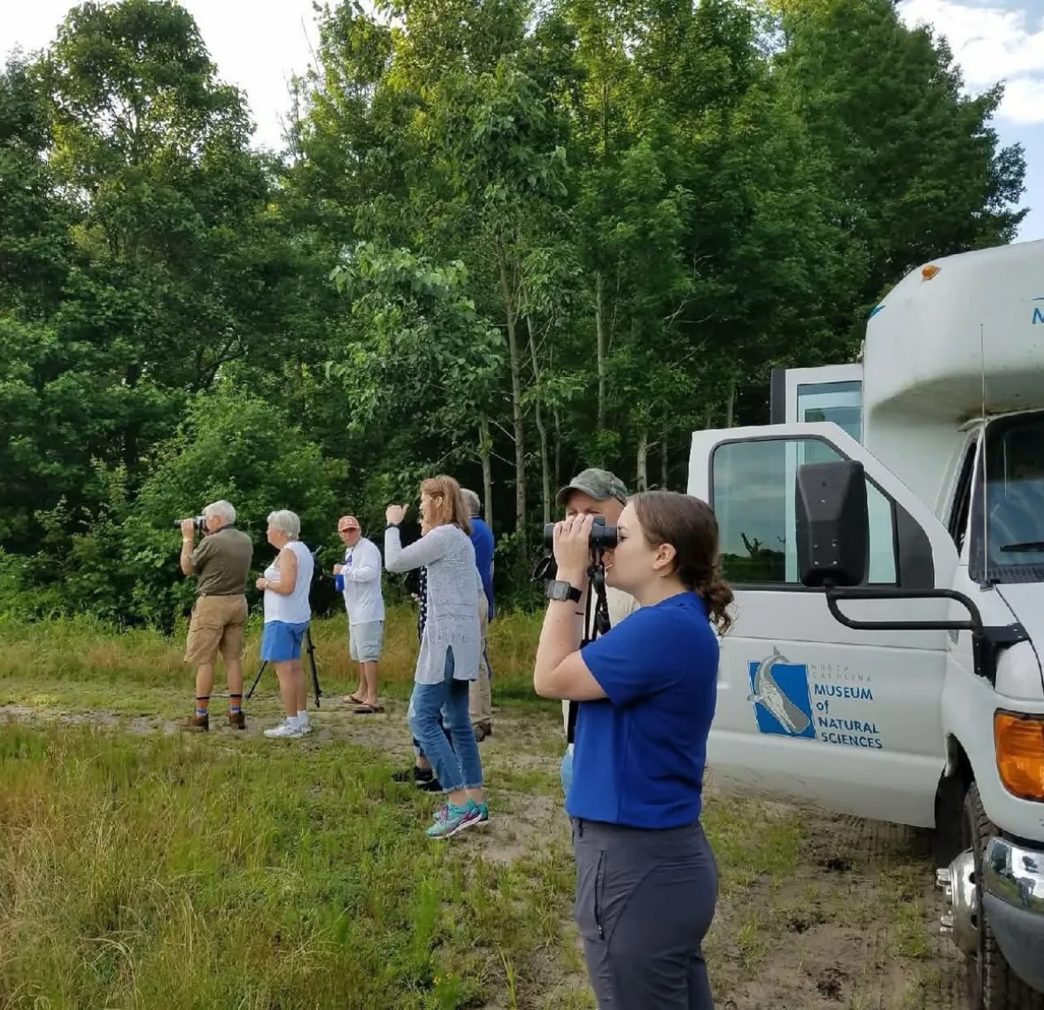 A group of people birdwatching near a white van in a grassy area by a forest.