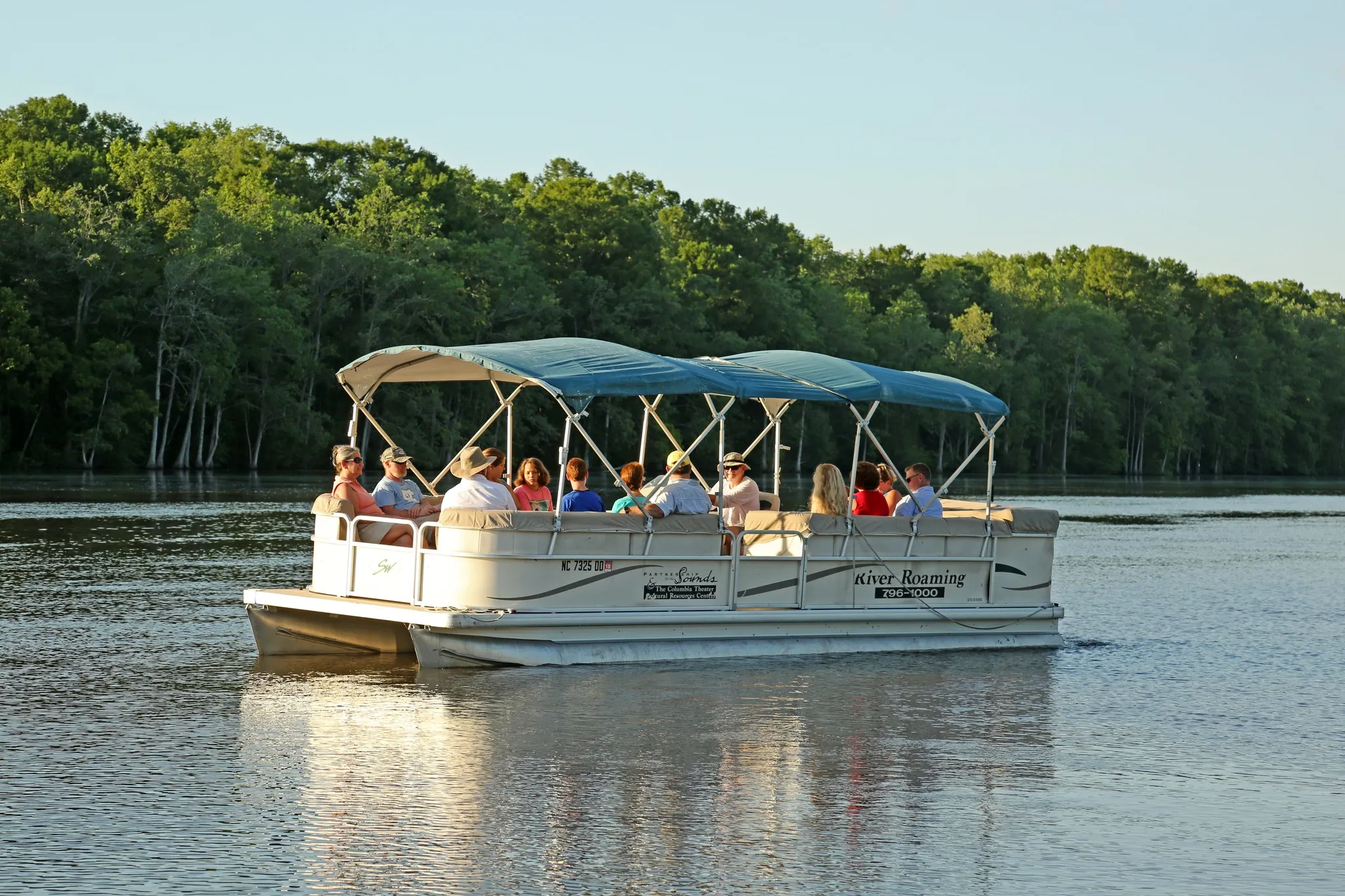 A pontoon boat with people on board cruises on a calm river with trees lining the shore in the background.