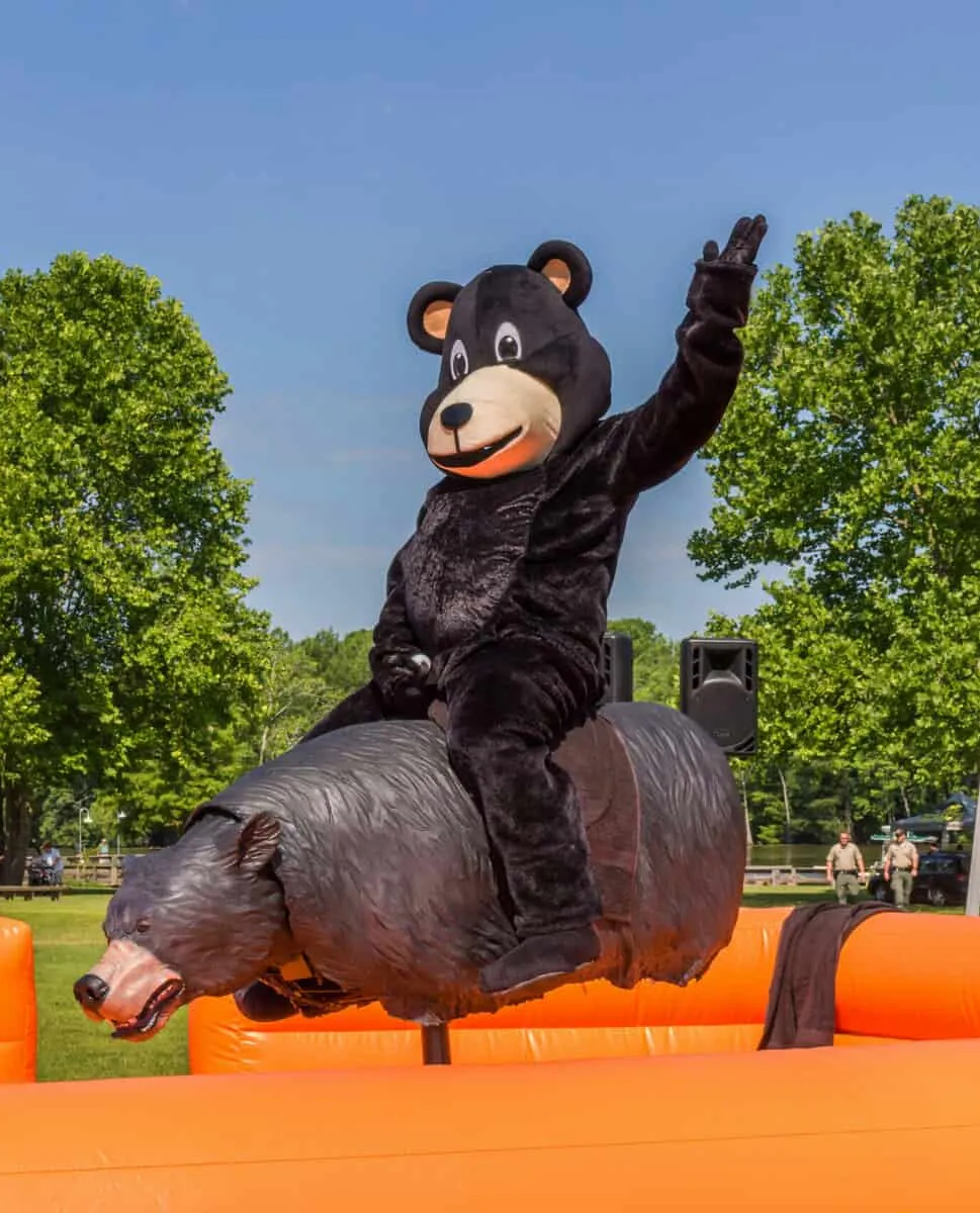 Person in a black bear costume rides a mechanical bear at an outdoor event with trees in the background.