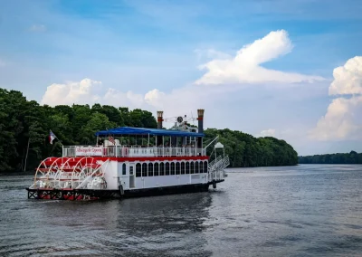 A white and red paddleboat cruises along a river with trees lining the water under a blue sky.
