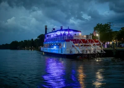 A riverboat with blue lights docks at night, reflecting on the water under a cloudy sky.