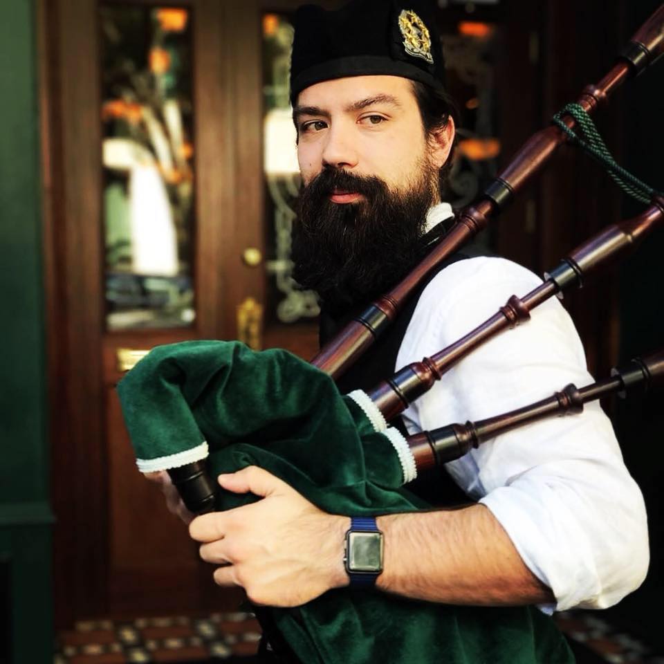A man with a beard plays the bagpipes, wearing a hat and a smart vest, standing by wooden doors.