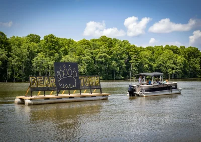 A boat pulls a floating "Bear Town" sign with a bear paw on a river, trees in the background.