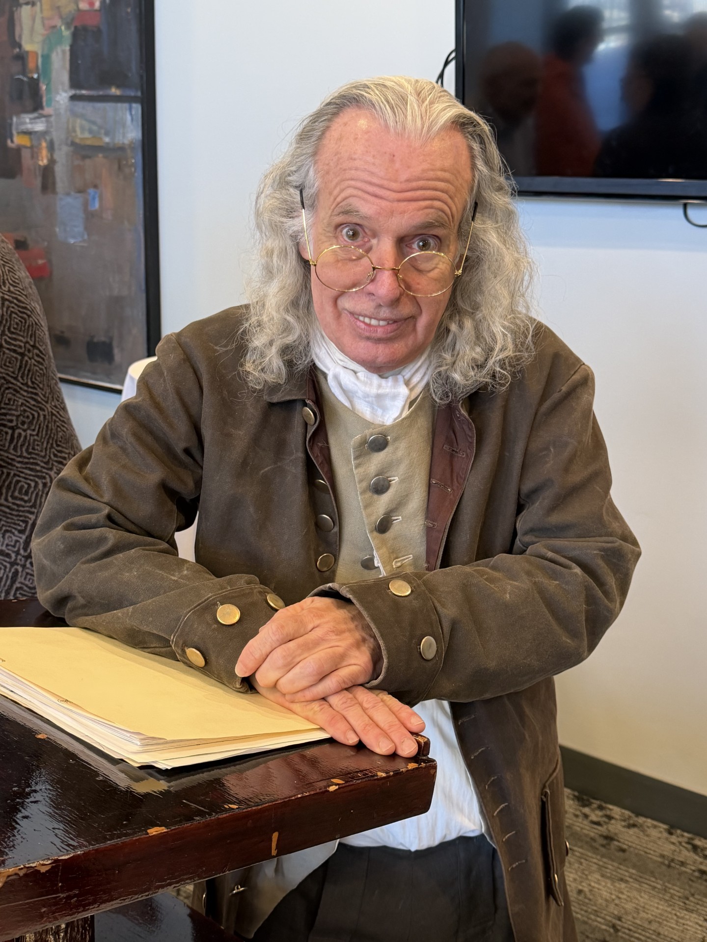 An older man in colonial attire with long gray hair and glasses sits at a table with papers, smiling.