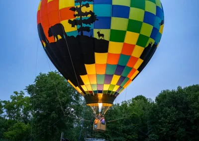 A colorful hot air balloon tethered to a pickup truck at a grassy field, with trees in the background.