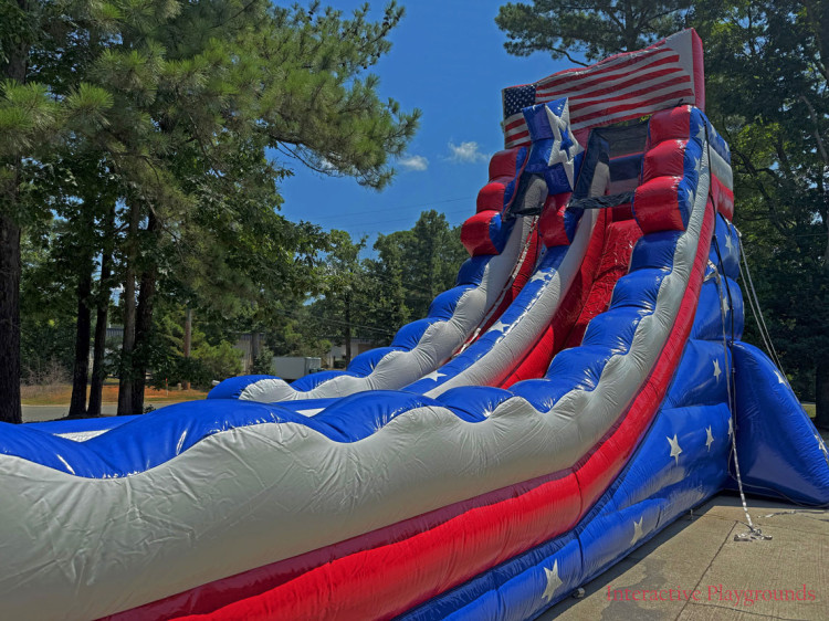 A tall inflatable slide with a stars and stripes American flag design, set outdoors near trees.