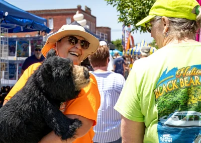 Smiling person holding a black bear plush at an outdoor festival, talking to another person in a bright shirt.