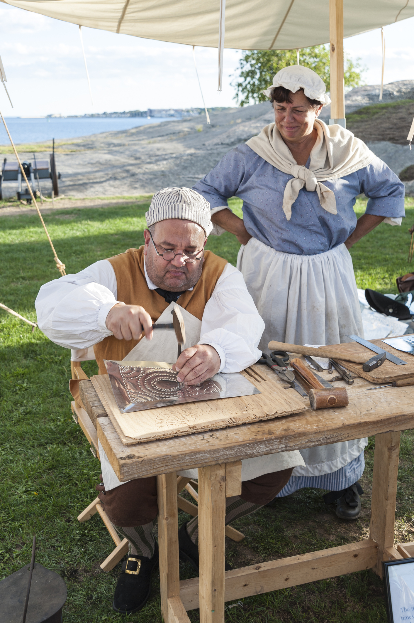 A man in historical clothing engraves metal as a woman in period dress stands beside him outdoors.