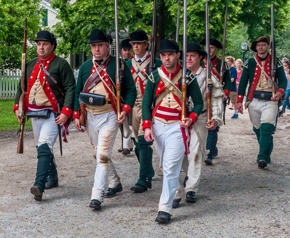 A group of reenactors in Revolutionary War-era uniforms march outdoors on a gravel path.