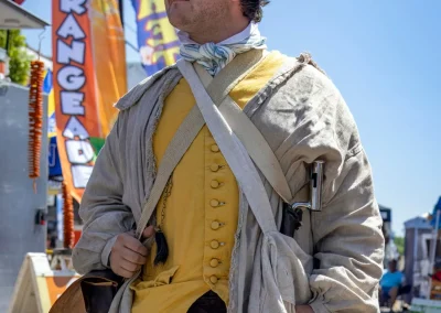 A man in colonial-era costume stands outdoors at a street fair, with colorful banners in the background.