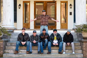 Six men, five seated on steps and one standing behind them, pose in front of a building with columns and glass doors.