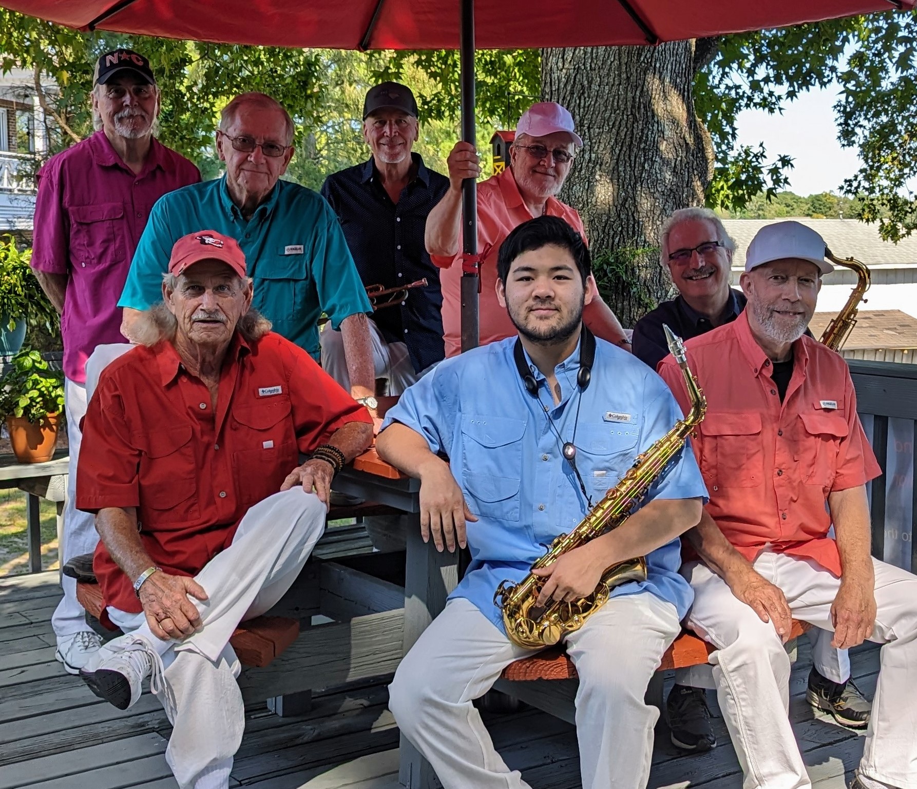 Eight men, some holding saxophones, sit and stand on a porch under a red umbrella, smiling at the camera.