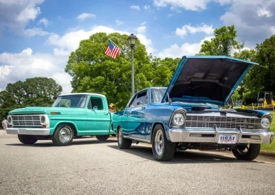 A teal vintage truck and a blue classic car with its hood open parked on a sunny street with an American flag.