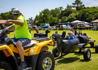 A man on an ATV pulls kids riding in barrel train carts at an outdoor event on a sunny day.