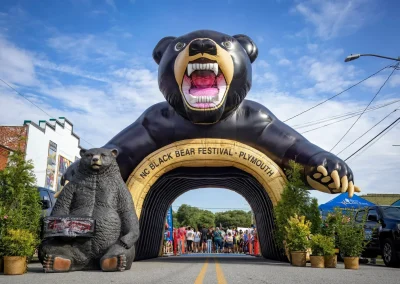 Large inflatable black bear arch at NC Black Bear Festival entrance, with crowds and a bear statue nearby.