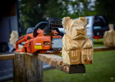 A wooden bear sculpture and a chainsaw rest on a wooden beam outdoors, with more carvings in the background.