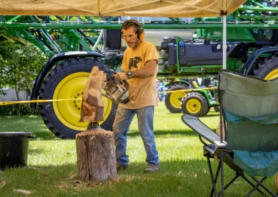 A man wearing ear protection uses a chainsaw on a log outdoors near green farm equipment.