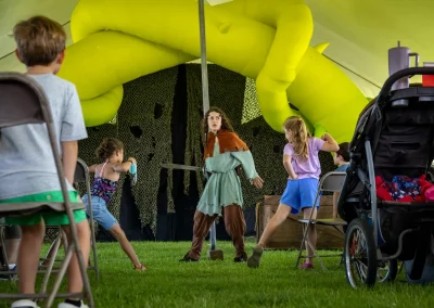 A performer in costume entertains children under a tent with inflatable yellow decorations and chairs.