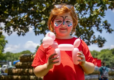 Child with face paint holds a balloon animal outdoors, wearing a red shirt and sunglasses on their head.