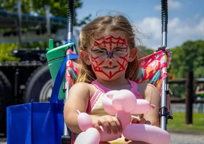 Smiling girl with a spiderweb face paint sits in a stroller holding a pink balloon animal outdoors.