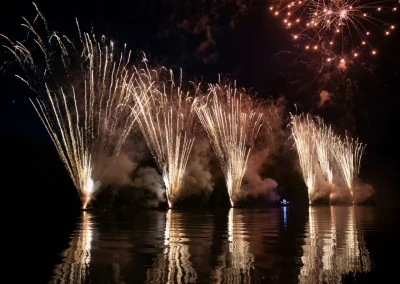 Golden fireworks burst and reflect over a calm lake at night, lighting up the dark sky and water.