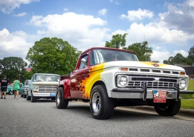 Vintage Ford trucks parked at a car show, with people walking and an American flag in the background.