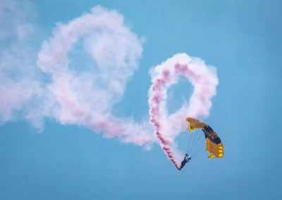 A skydiver with a yellow parachute makes a heart-shaped smoke trail against a blue sky.