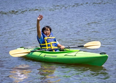 Person in a green kayak wearing a life jacket, waving while paddling on a calm body of water.