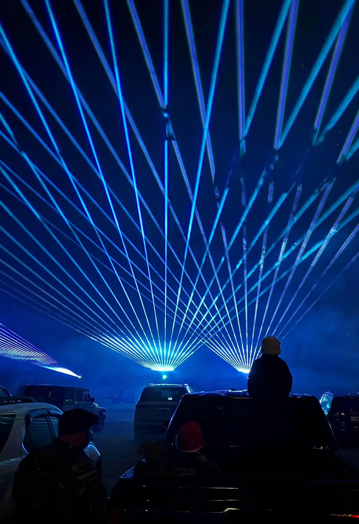 People watch blue laser lights in the night sky from parked cars at an outdoor event.