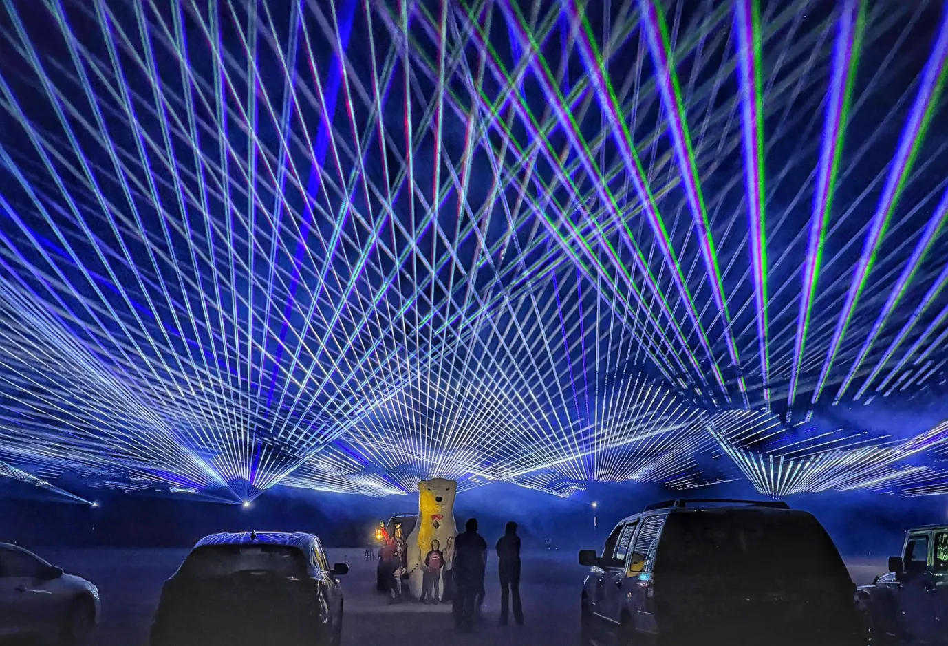 People and cars under a night sky filled with crisscrossing blue and green laser lights.