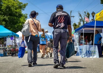 People walk through an outdoor market on a sunny day, carrying bags and wearing casual clothing and hats.