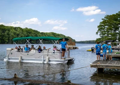 A group rides a pontoon boat on a lake while kids in blue shirts stand on a dock under a sunny sky.