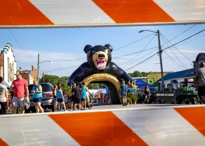 People gather at a festival entrance with a large inflatable black bear, seen through an orange-striped barrier.