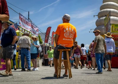 A man in an orange shirt sits on a stool at a busy outdoor fair with food stands and crowds.