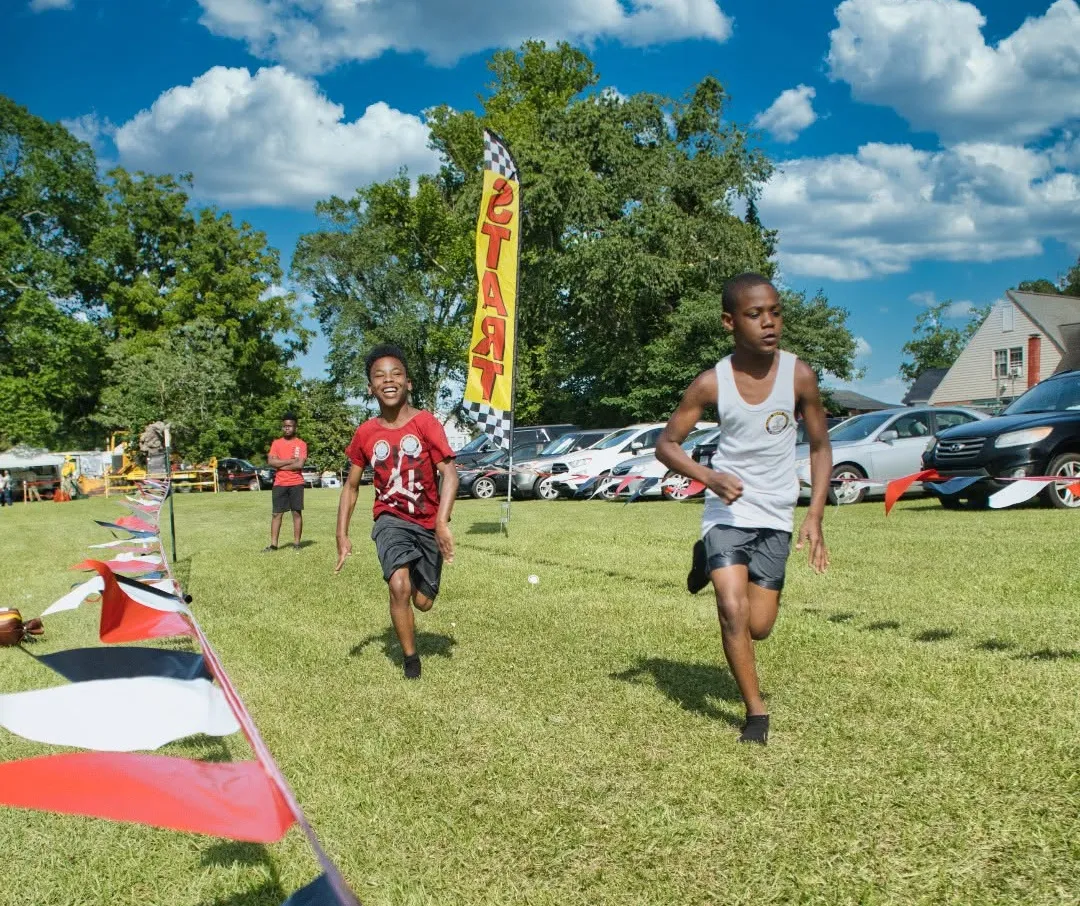 outrun-1 Two boys race toward a finish line at an outdoor event, with a "START" flag and cars in the background.